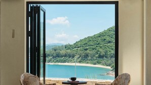 Una habitación con un gran ventanal arqueado, sillas de mimbre, una mesita y vistas a una playa y montañas.