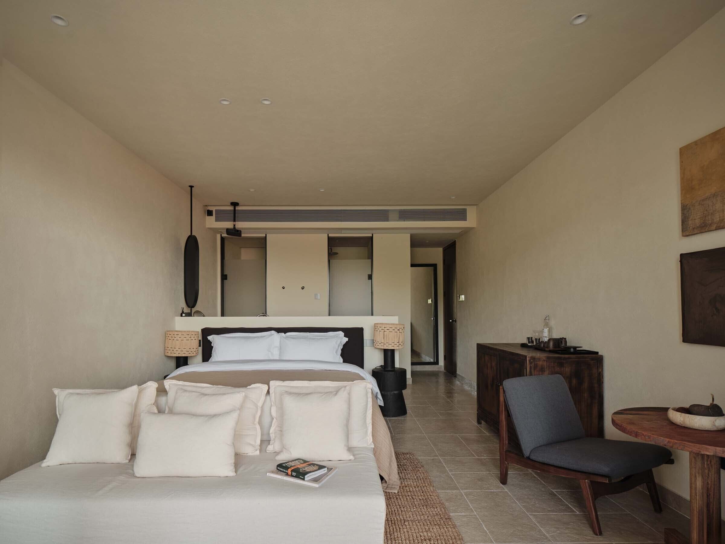 Habitación de hotel con cama, lámparas de noche, una silla, una mesita y vistas al pasillo.