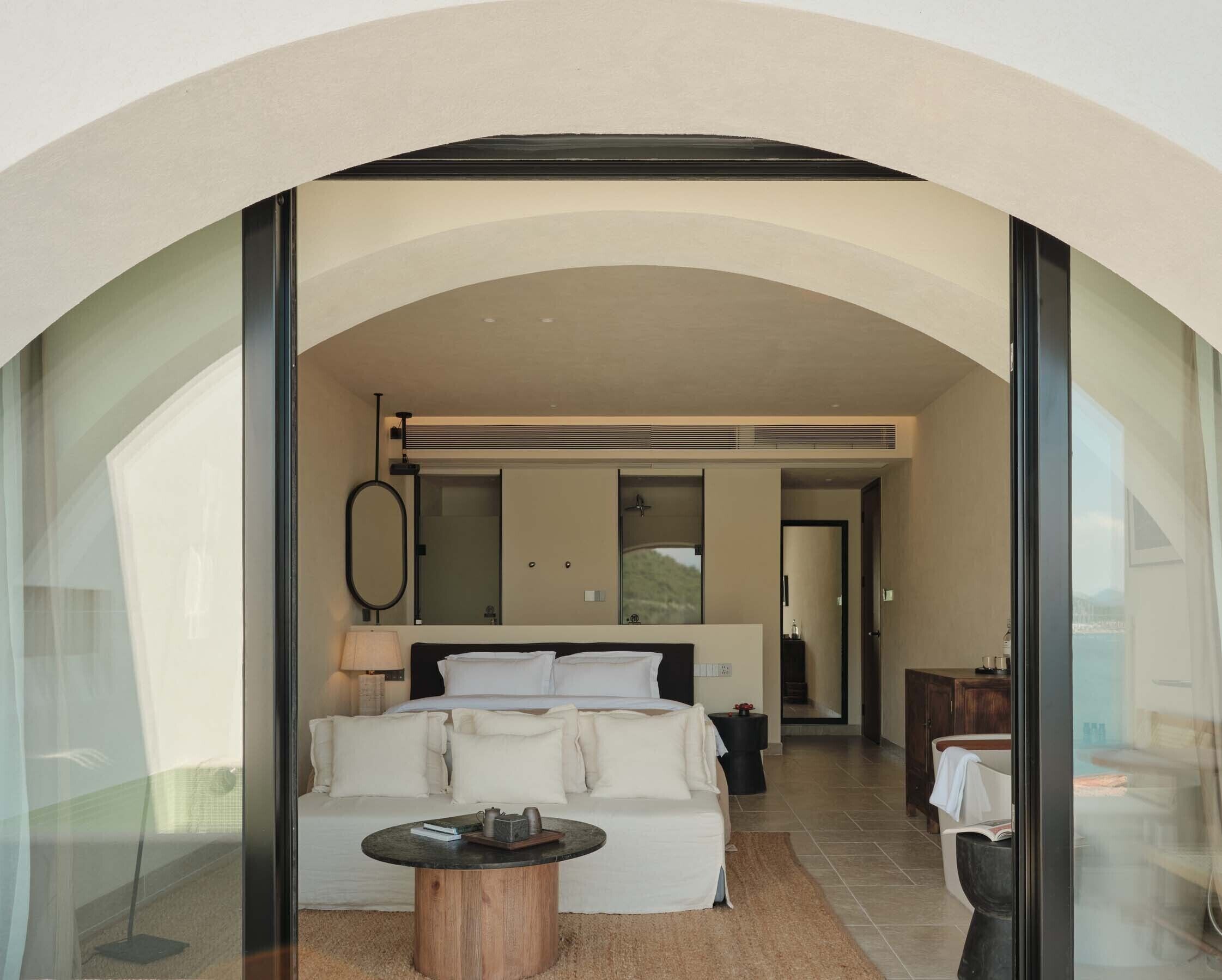 Un dormitorio moderno con una cama grande, una mesa de centro redonda de madera y vistas a través de una puerta de cristal.