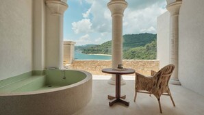 Un área de spa con una bañera de hidromasaje circular, una mesita redonda y una silla de mimbre, con vistas panorámicas a una playa y montañas.
