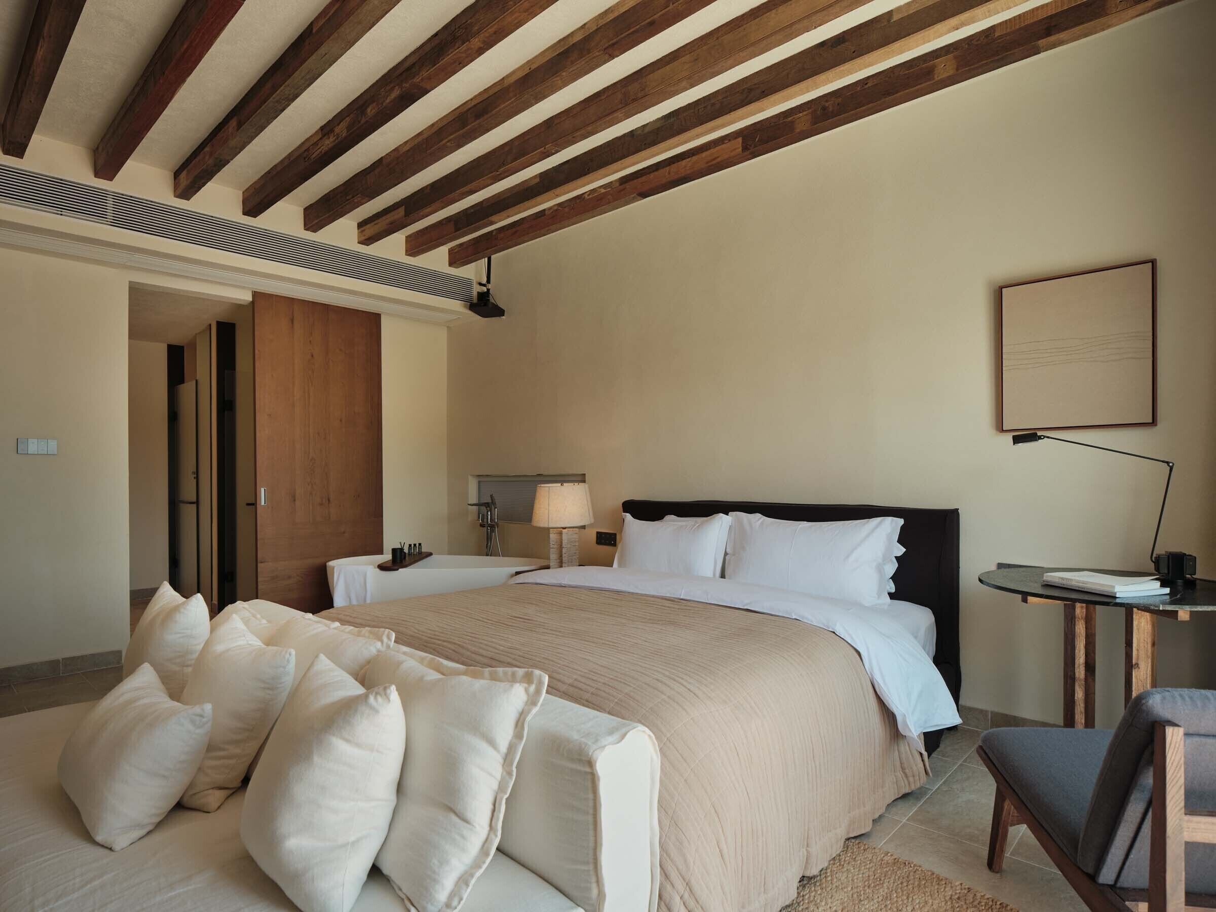 A bedroom with a large bed, wooden beams on the ceiling, a bedside table with a lamp, and a chair.