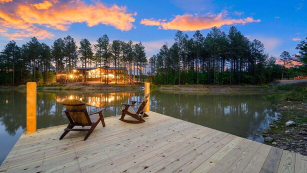 Un muelle de madera con dos sillas Adirondack, un lago y una casa con luces que se reflejan en el agua.
