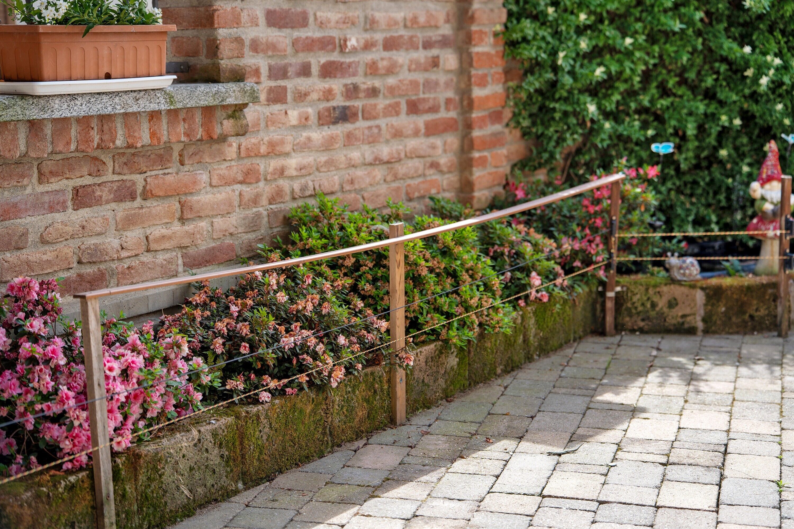 Un mur de briques avec une rambarde en bois, un chemin de pierres et des plantes en fleurs.