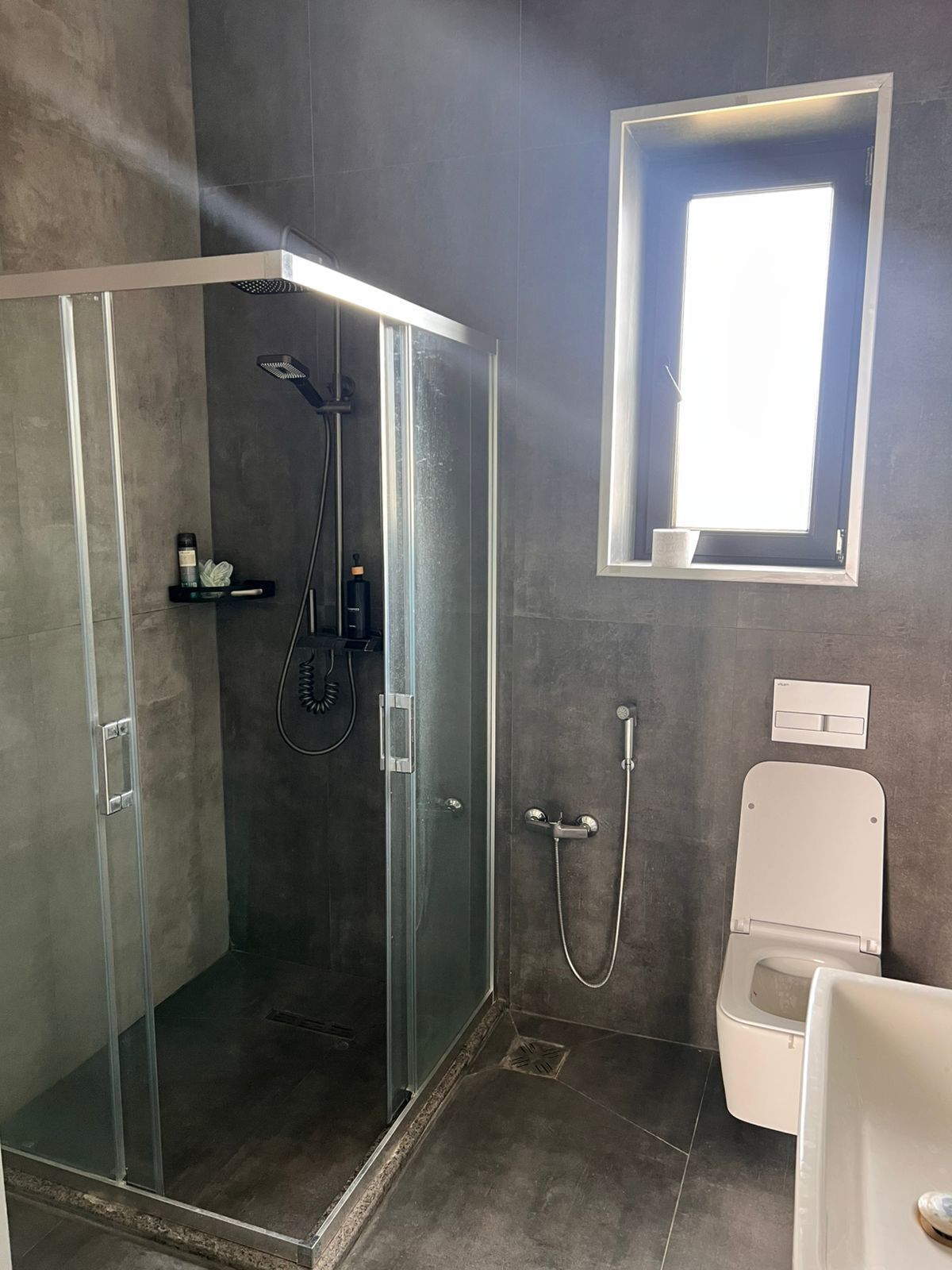 A modern bathroom with a glass shower enclosure, a toilet, and a window above the toilet.