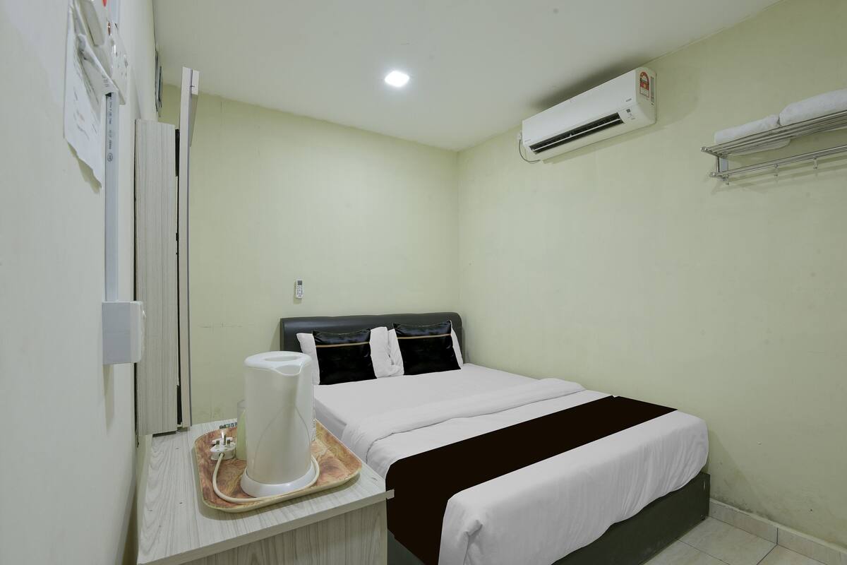 Una habitación de hotel con una cama, una mesita de noche con hervidor, un equipo de aire acondicionado y un perchero en la pared.