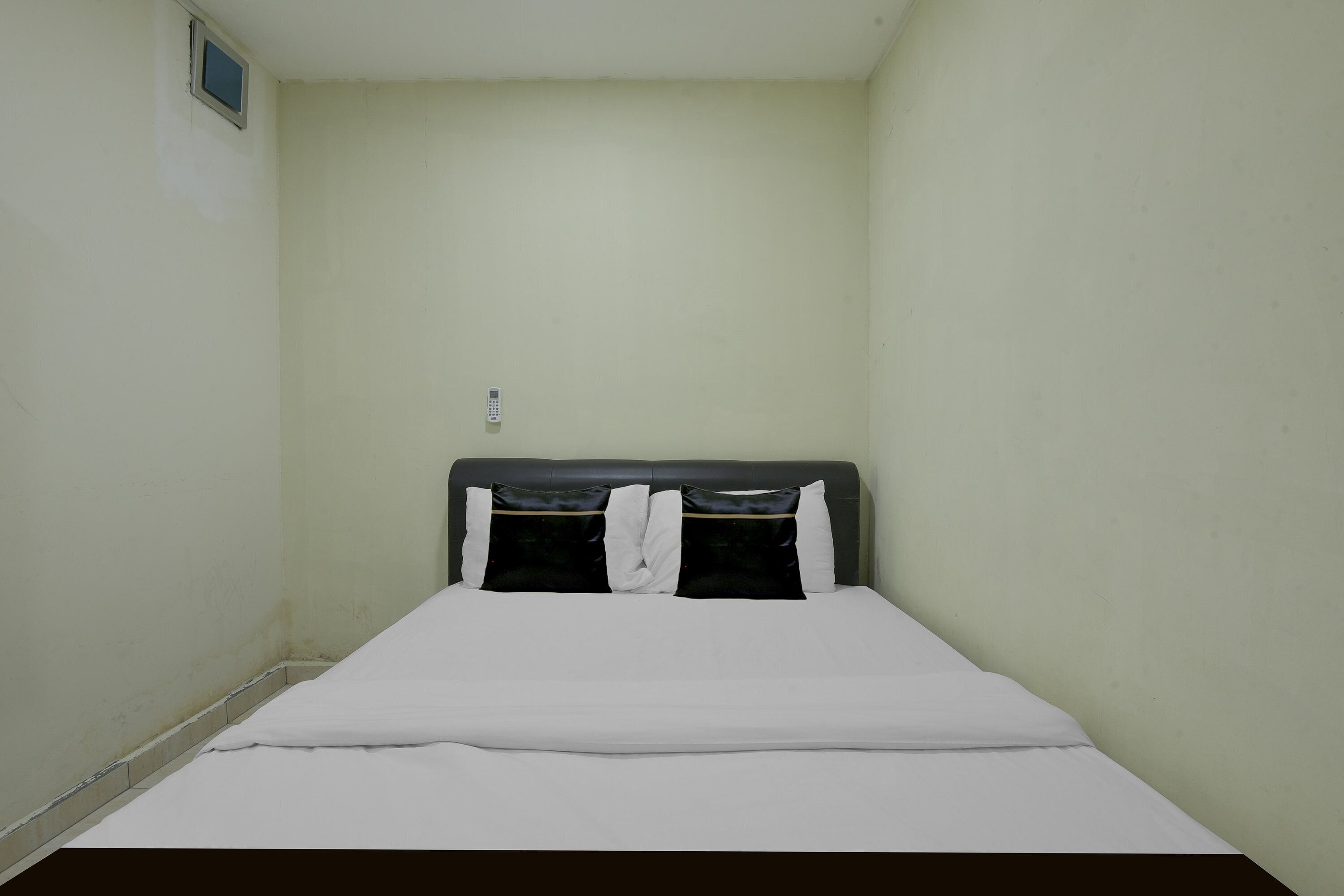 A small, single-bed room with a plain headboard, a single wall-mounted light, and a small window.