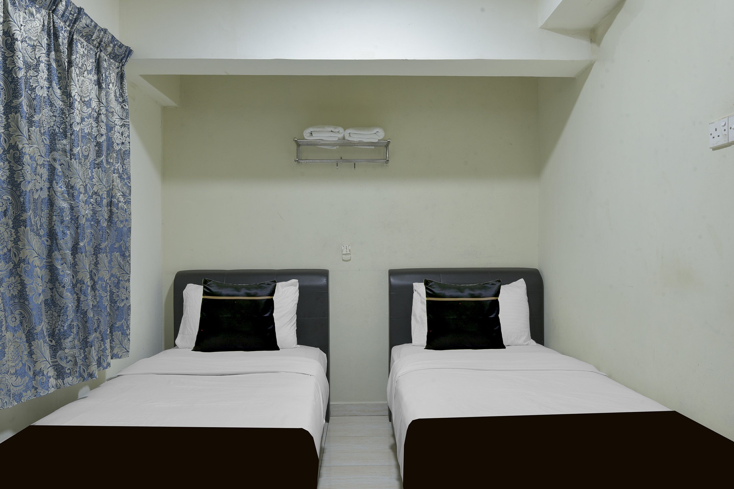 A room with two beds, a curtain, a shelf with towels, and a light switch.