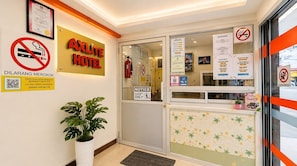 Entrada de un hotel con un letrero que dice 'AXLITE HOTEL', un cartel de 'no fumar', un aviso y un código QR para información.