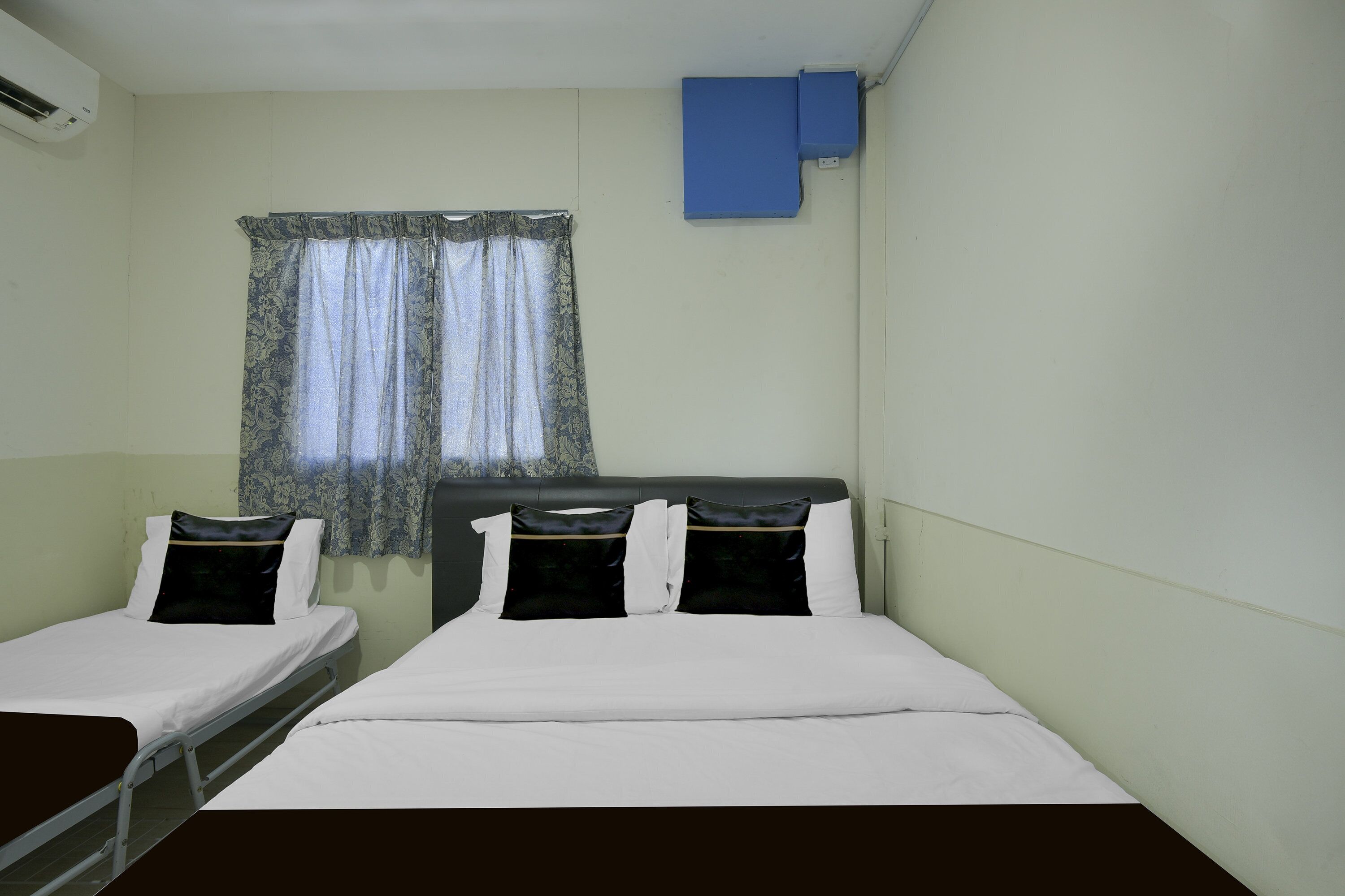 A room with two beds, a window with curtains, a wall-mounted air conditioner, and a blue object on the wall.