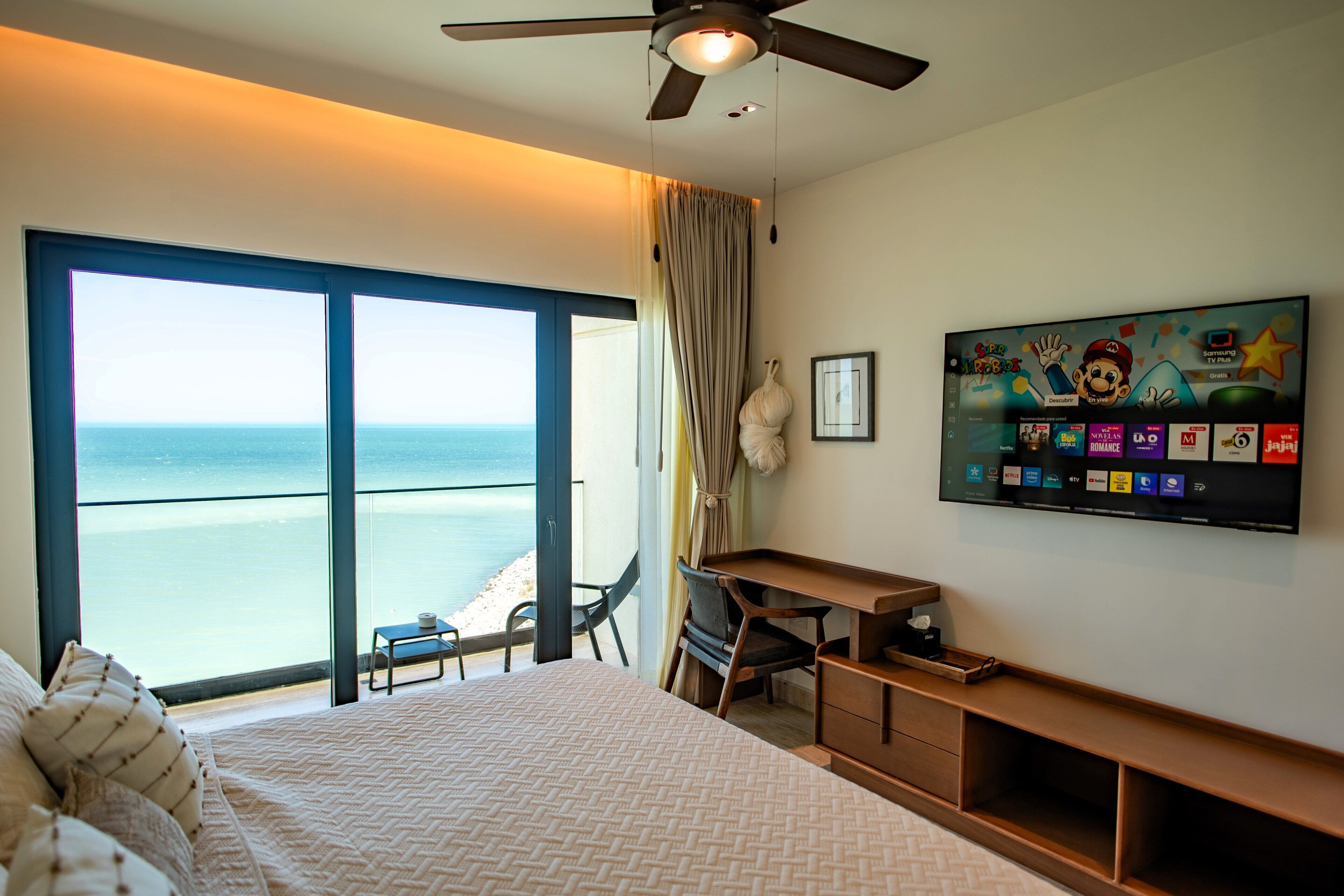 Quarto de hotel com cama, escrivaninha, cadeira, televisão e vista para o oceano.