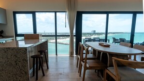 Cozinha moderna com bancada de granito e banquetas de madeira, área de refeições com mesa e cadeiras de madeira, e grandes janelas com vista para o oceano e praia.