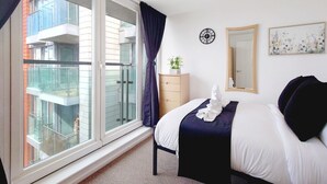 A bedroom with a large window, a bed, a dresser, a mirror, and a clock on the wall.