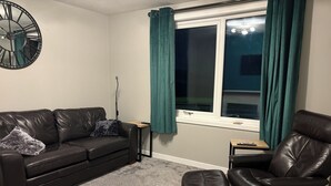 A living room with a leather sofa, ottoman, and chair, a clock on the wall, and a window with curtains.