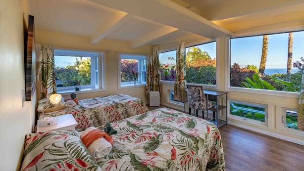 Une chambre avec deux lits, un bureau et de grandes fenêtres offrant une vue sur la mer et la végétation.