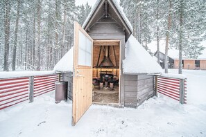 Una cabaña de madera con techo inclinado, una puerta abierta a un cuarto con chimenea y un bosque nevado al fondo.