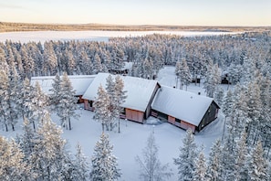 A lodge with multiple buildings surrounded by a dense forest, a frozen lake in the background, and snow-covered trees. - Wellevi 1 by Aavalevi (Levi)