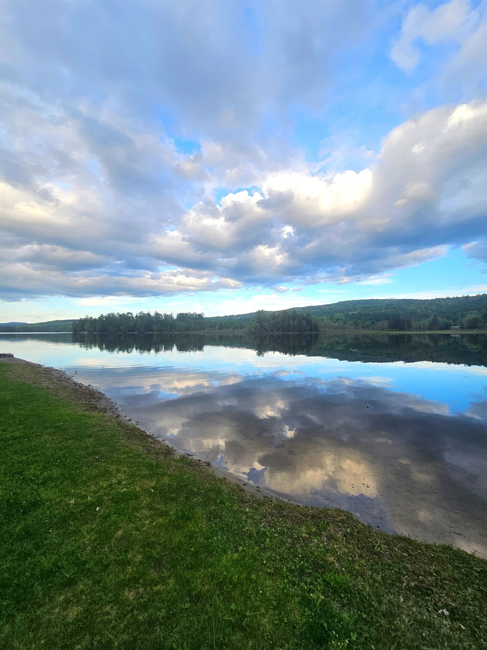 Ein ruhiger See mit einer grasbewachsenen Uferlinie und einem bewaldeten Hügel im Hintergrund.