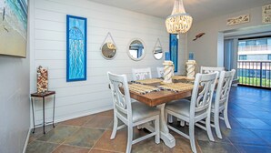 A dining area with a wooden table, white chairs, a chandelier, and wall art including a beach scene and mirrors. - Bayfront Sanibel Condo Water Views Pool Mariner Pointe 712 Dolphins Reach (Sanibel Island)