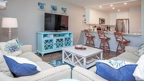 A living room with a TV, blue cabinet, and a kitchen area with bar stools. - Bayfront Sanibel Condo Water Views Pool Mariner Pointe 712 Dolphins Reach (Sanibel Island)