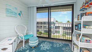 A balcony with a white wicker chair, a blue patterned ottoman, a white bookshelf with books, and a view of a building and greenery. - Bayfront Sanibel Condo Water Views Pool Mariner Pointe 712 Dolphins Reach (Sanibel Island)