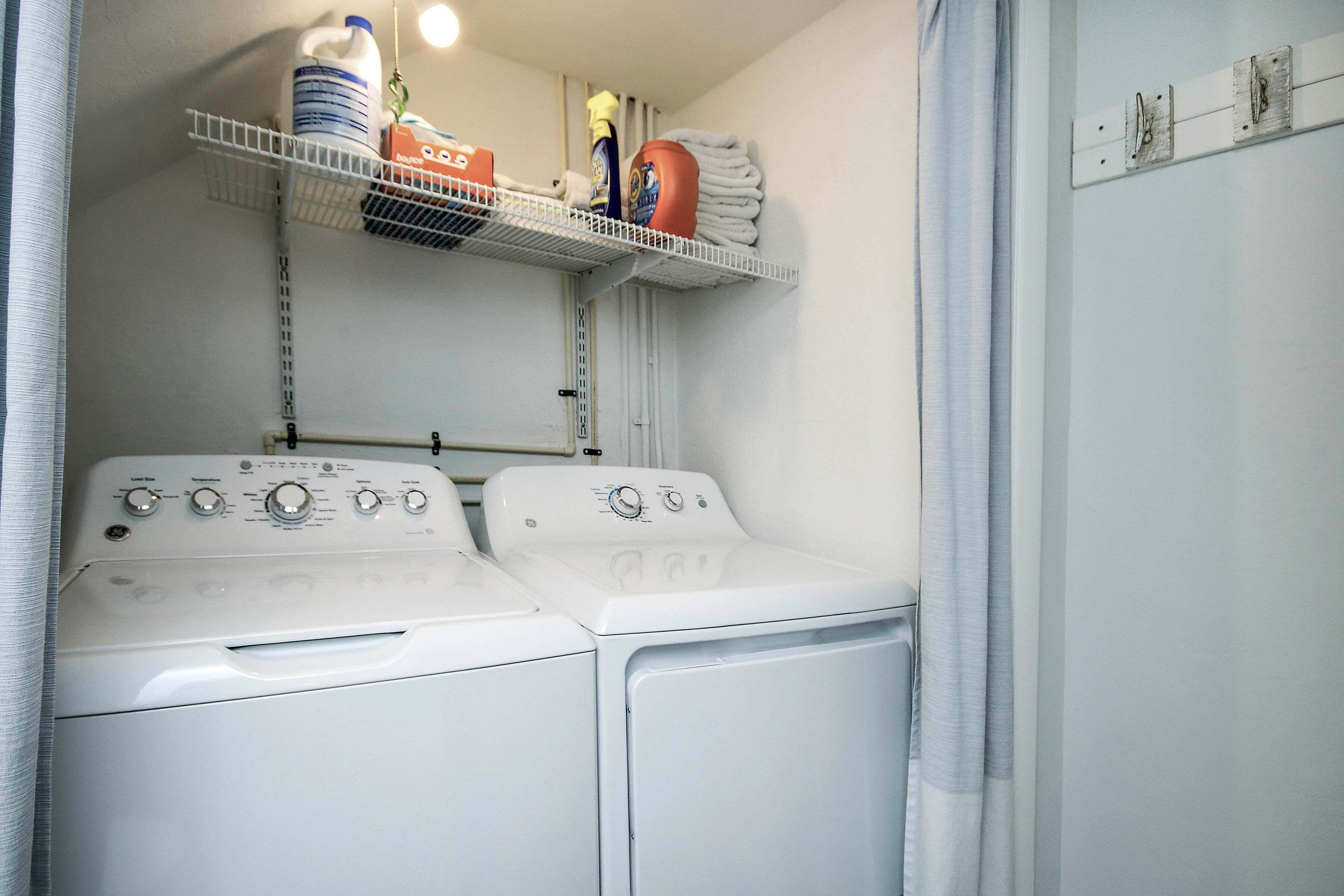 A laundry area with two white washers and dryers, a metal shelf with laundry supplies, and a white curtain.