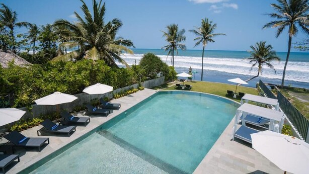 Une piscine entourée de chaises longues et de parasols, bordée de palmiers et offrant une vue sur l’océan.