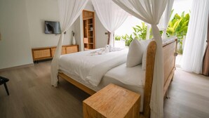 A bedroom with a four-poster canopy bed, a wooden bedside table, a TV mounted on the wall, and a view of greenery outside. - Nyuh Lengkong Beachfront Villa (Tabanan)