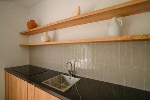 A kitchen with a black countertop, a stainless steel sink, wooden shelves, and white tiled backsplash. - Nyuh Lengkong Beachfront Villa (Tabanan)