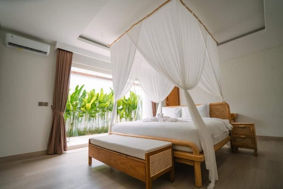 Un dormitorio con cama con dosel, un banco de madera, una mesita de noche y vistas a zonas verdes al exterior.