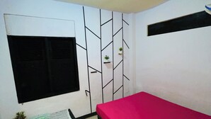 A bedroom with a bed, a window, a wall-mounted shelf with plants, a small table, and a floor mat.
