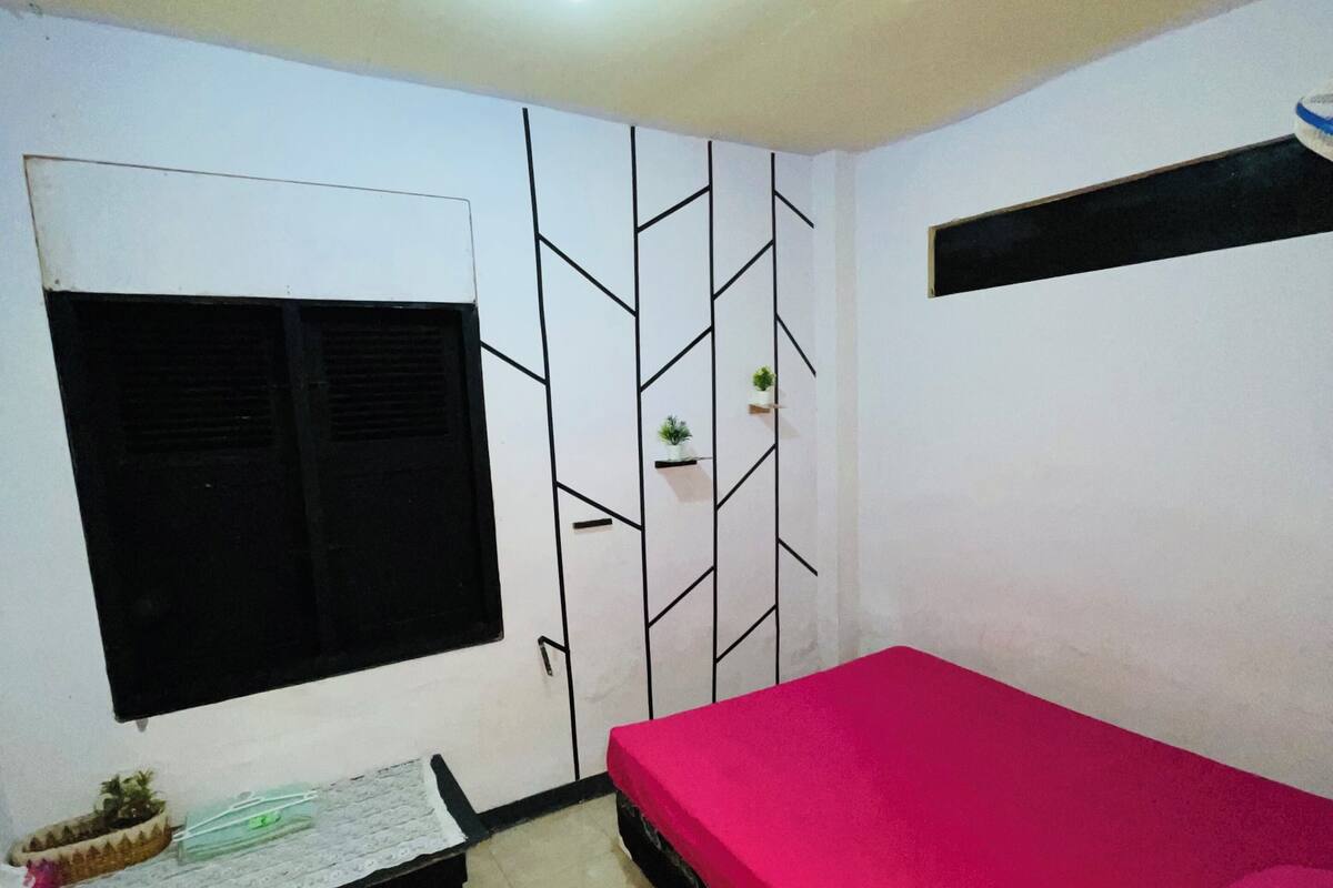 A bedroom with a bed, a window, a wall-mounted shelf with plants, a small table, and a floor mat.