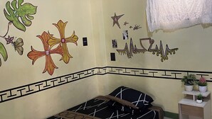 A room with a bed, a nightstand, a window with lace curtains, and wall decorations including a star and a heart.