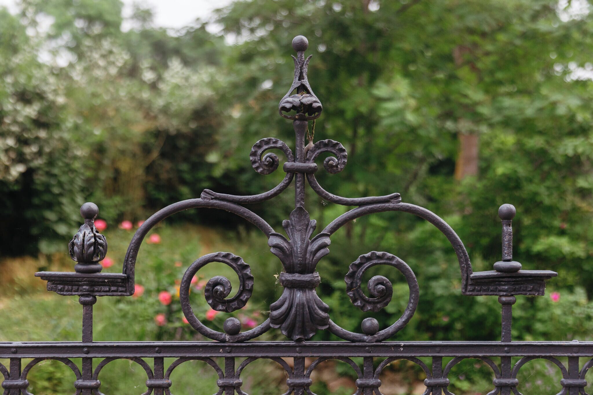 Una valla de hierro forjado con elaborados adornos en voluta y remates, frente a un exuberante jardín.