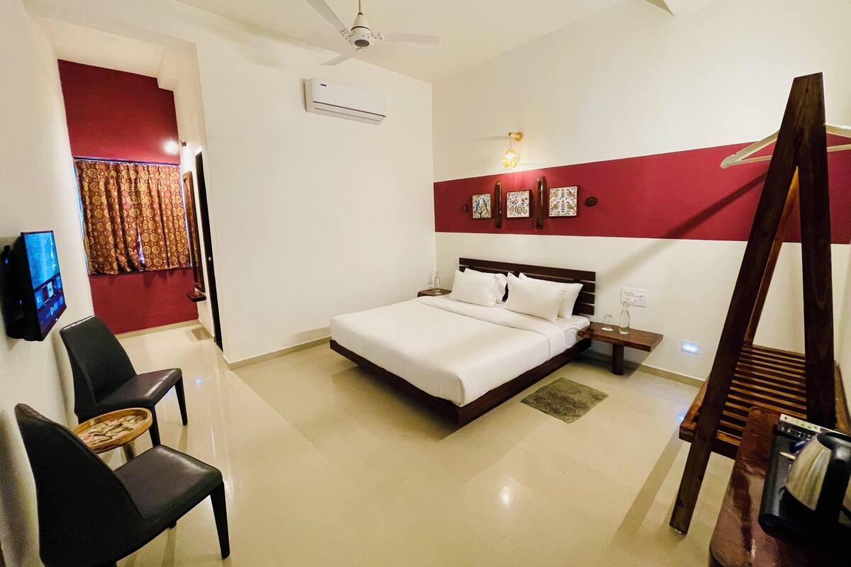 A hotel room with a bed, a TV, a ceiling fan, an air conditioning unit, a desk with a chair, and a red accent wall.