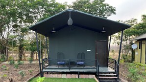 Un pequeño edificio con fachada oscura, un área de estar al aire libre techada con dos sillas, una puerta y una antena parabólica.