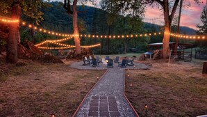 Una terraza que conduce a un área de estar con sillas y una chimenea, rodeada de árboles y luces colgantes.