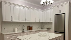 Una cocina con armarios blancos, encimera de mármol, refrigerador de acero inoxidable y una araña.
