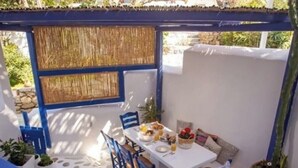 Un espace extérieur abrité, aménagé pour les repas, avec une table blanche dressée pour deux personnes, entourée de chaises bleues, de plantes en pot et d’un sol dallé imitant la pierre.
