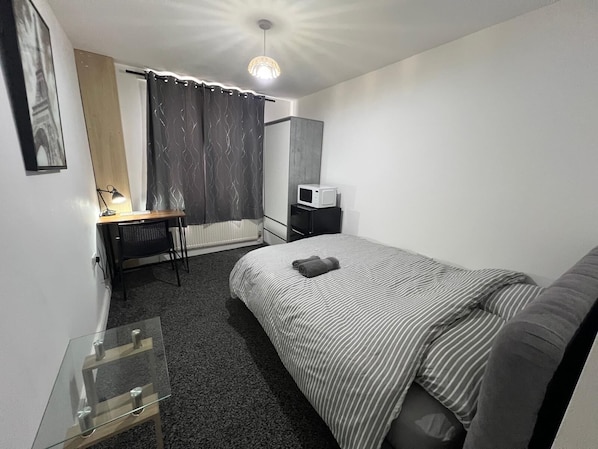 A hotel room with a bed, a desk with a lamp, a microwave, and a window with curtains.
