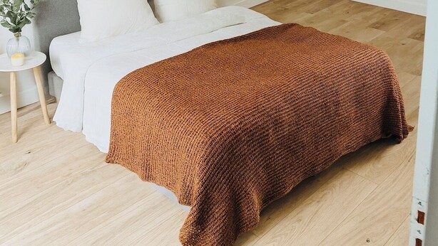 Une chambre à coucher avec un lit, une tête de lit grise, une plante décorative dans un cadre, une petite table d’appoint et un plancher en bois.