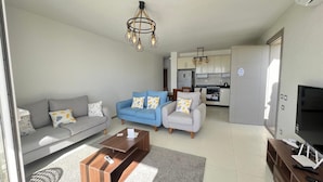 A modern living room with a sofa, armchairs, a coffee table, a TV, and a kitchen in the background.