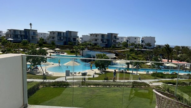 A pool area with multiple swimming pools, a grassy lawn, and modern buildings in the background.