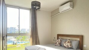 A bedroom with a bed, a nightstand, an air conditioning unit, a window with a view of a pool and greenery, and a hanging light fixture.