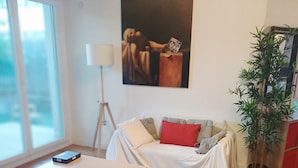 En stue med trægulv, en hvid sofa med røde puder, et hvidt bord, et billede på væggen og en potteplante.