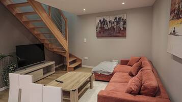 Un salon avec un téléviseur, une table basse en bois, un canapé rouge, un escalier et un tableau représentant un paysage urbain.