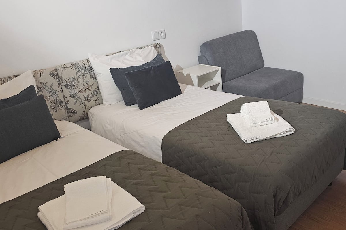 A hotel room with two beds, a chair, a small table, and a trash bin.