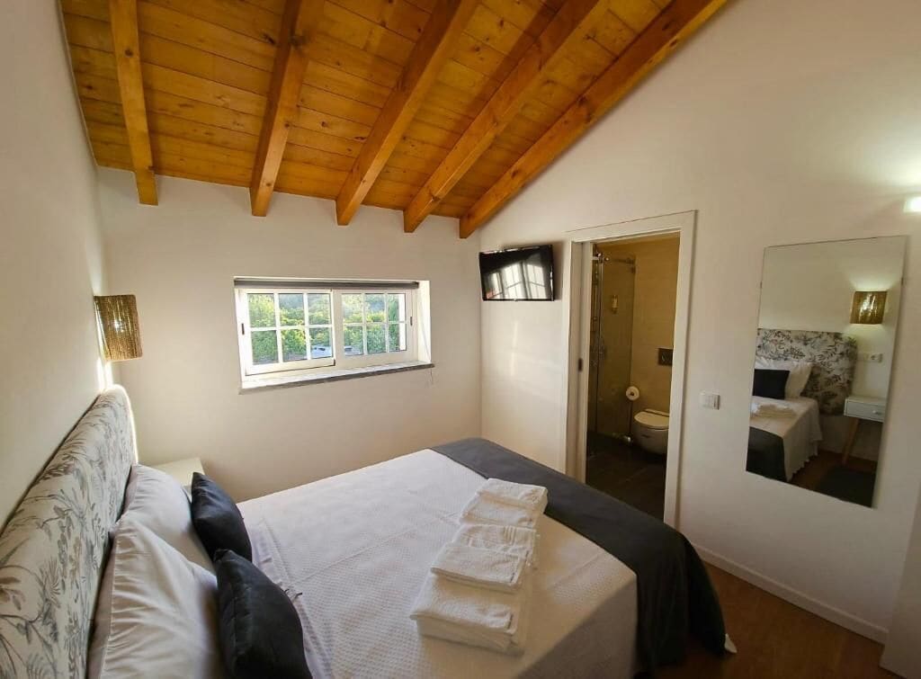 Een slaapkamer met een houten plafond, een bed met witte lakens, een raam, een deur naar een badkamer, en een spiegel aan de muur.