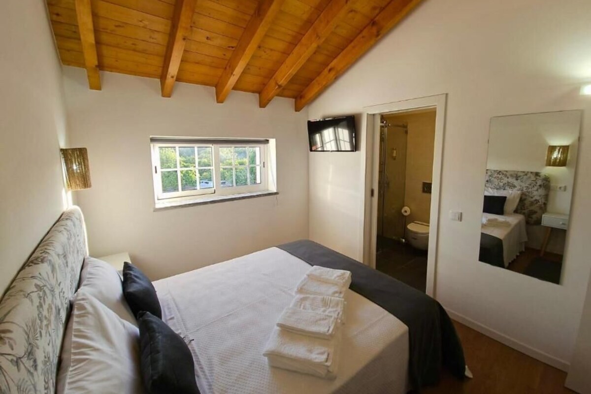 Een slaapkamer met een houten plafond, een bed met witte lakens, een raam, een deur naar een badkamer, en een spiegel aan de muur.