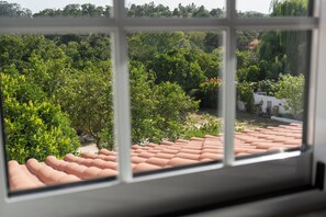 Vista pela janela, com telhado de azulejos, vegetação e um edifício branco ao fundo.