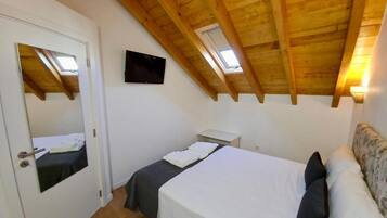 A bedroom with a sloped wooden ceiling, a bed with white linens, a small bedside table, a mirror, and a TV mounted on the wall.